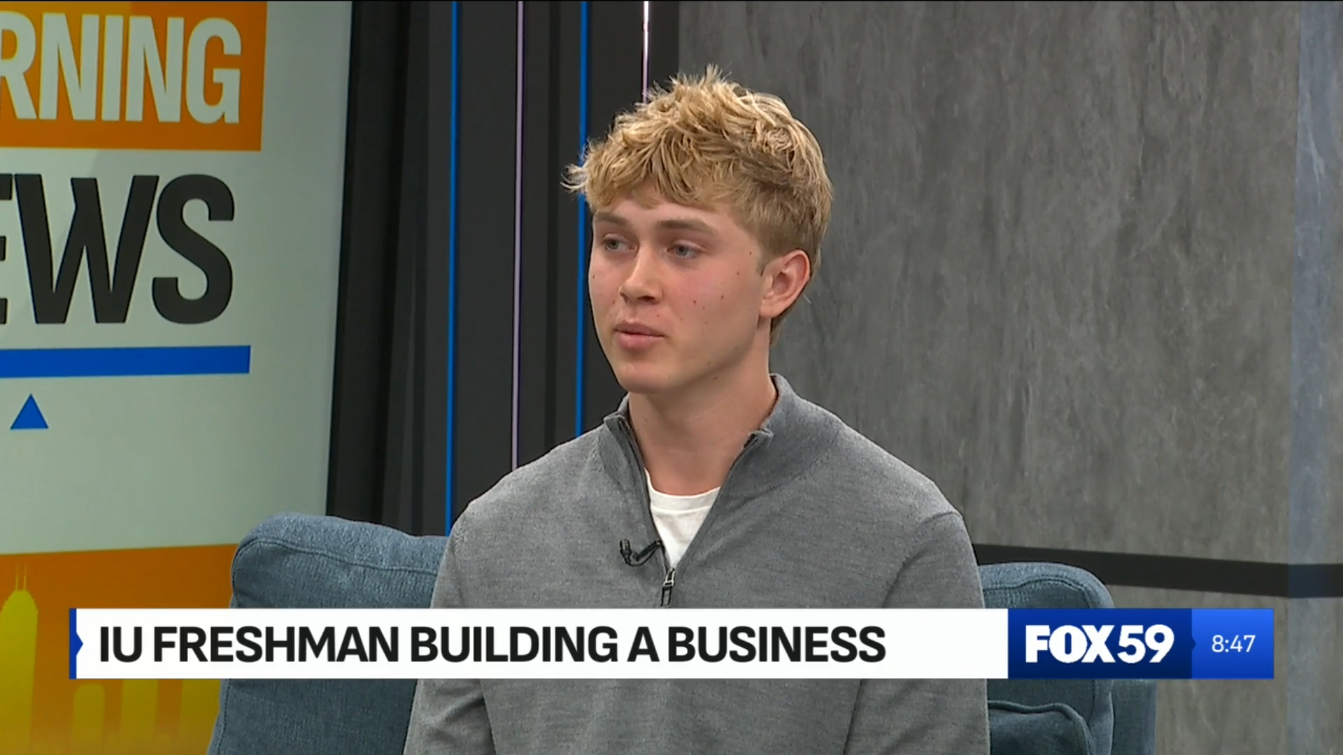 Young man in a gray sweater sitting on a blue couch with a 'FOX 59' news banner in the background.